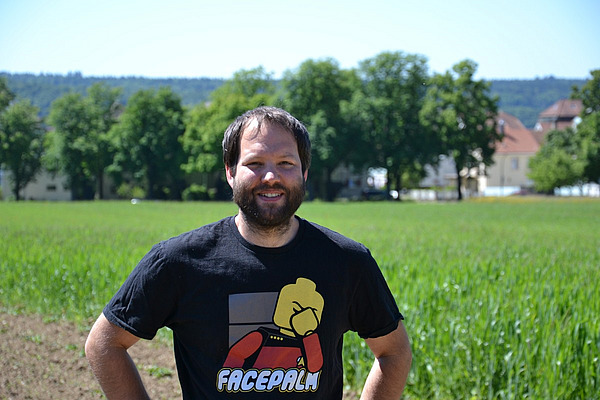 Ein Mann mit Bart steht vor einem Feld, dahinter einige Bäume.