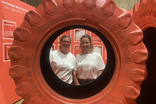 Zwei Frauen stehen hinter einem rot angemalten Traktorrad und schauen durch die Öffnung.