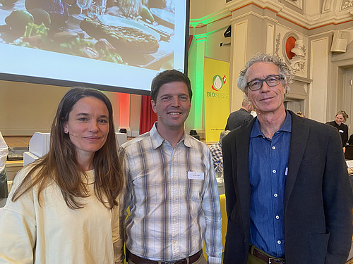 Eine Frau und zwei Männer posieren in einem üppig dekorierten Saal. 