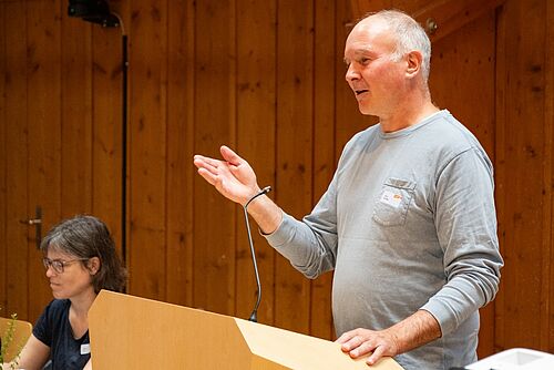 Ein Mann referiert an einem Rednerpult stehend, links davon eine Frau sitzend.