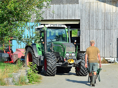 Ein Bauer läuft mit einer Schubkarre und dem Rücken zur Kamera Richtung Traktor und Scheune