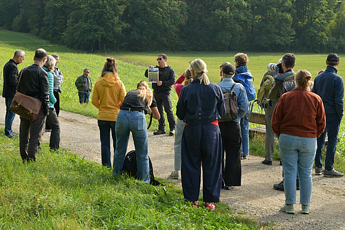 Eine Gruppe Menschen steht auf einem Weg zwischen zwei Grünflächen. Ein Mann in der Mitte zeigt ein Foto von einer blühenden Fläche. 