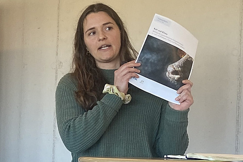 Eine Frau hält eine Broschüre, im Hintergrund eine Präsentation auf einem Bildschirm. 