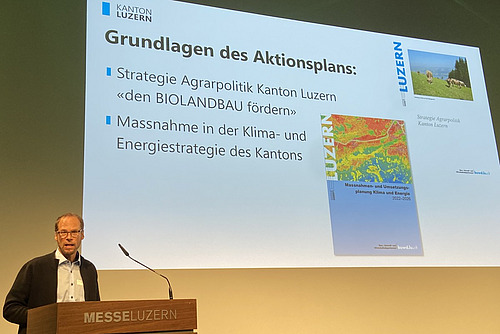 Ein Mann referiert an einem Pult vor einer Powerpoint-Präsentation. 