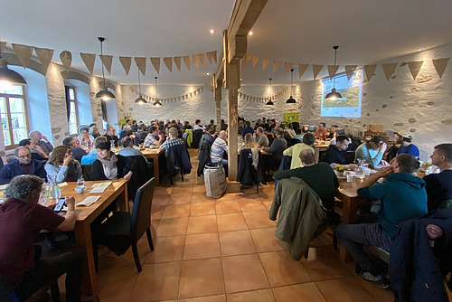 Blick in einen gut gefüllten Saal mit Holzbalken, wo Dutzende von Menschen an Tischen sitzen. 