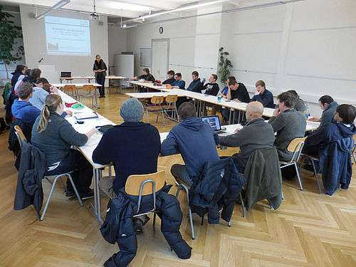 Eine Gruppe von Menschen sitzen in einem Raum, eine Frau hält eine Präsentation.