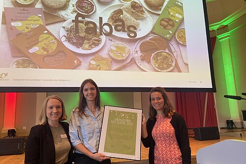 Drei Frauen vor einer Leinwand mit einer Auszeichnung.