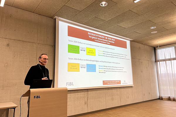 Eine Person hält einen Vortrag, im Hintergrund sind die Folien der Powerpoint-Präsentation zu sehen.