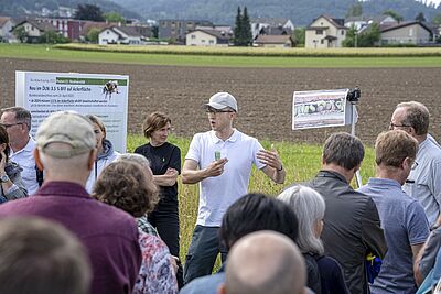 Ein Referent gestikuliert an einem Feldrand vor einer Gruppe Zuhörerinnen und Zuhörer.