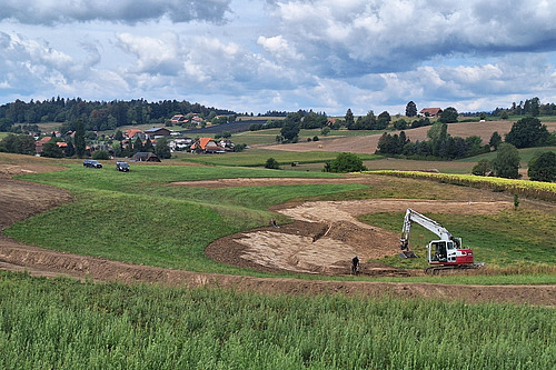 Zu sehen ist ein landwirtschaftlich geprägtes Tal. Auf den Flächen im Vordergrund werden mit einem Bagger grossflächige Erdarbeiten durchgeführt.