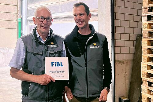 Zwei Männer lächeln und halten die FiBL-Plakette in der Hand