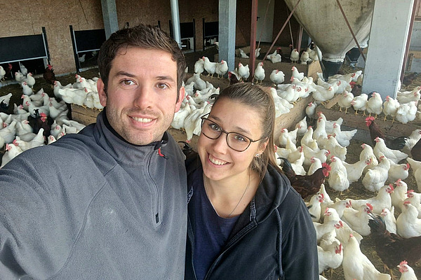 Selfie-Aufnahme von einem Mann und einer Frau mit Hühnern im Hintergrund.