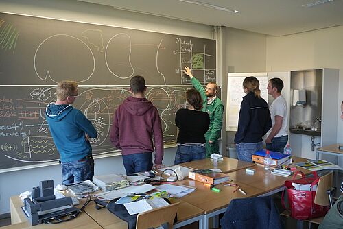 Lehrperson zeigt Studierenden eine Zeichnung auf der Wandtafel.