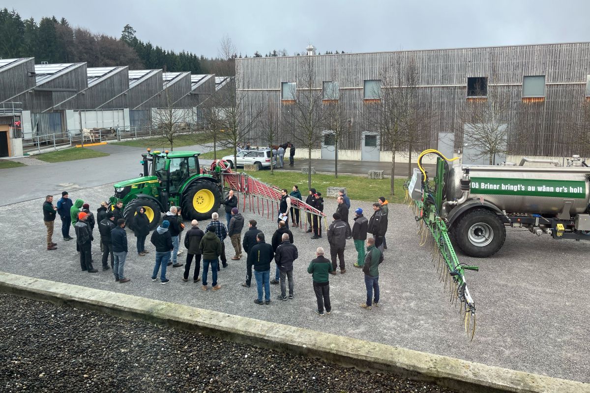 Menschen stehen auf einem Platz und betrachten einen Traktor mit Schleppschlauch und ein Güllefass.